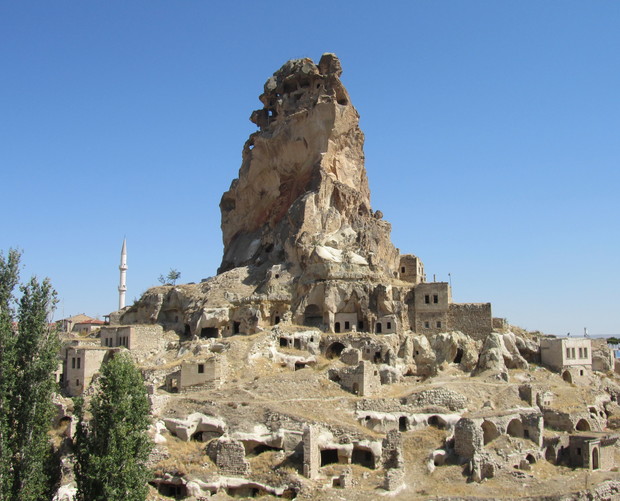 Rock formation stands tall with carved cavities and nearby ancient dwellings, set in an arid landscape under a clear blue sky, accompanied by a distant minaret.